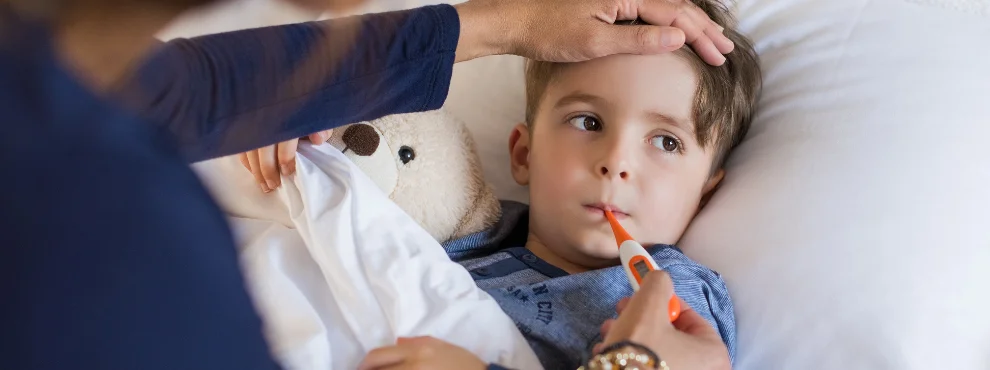 A toddler is lying in bed with a thermometer in their mouth. A woman, probably the mother, is placing a hand on the child’s forehead. 