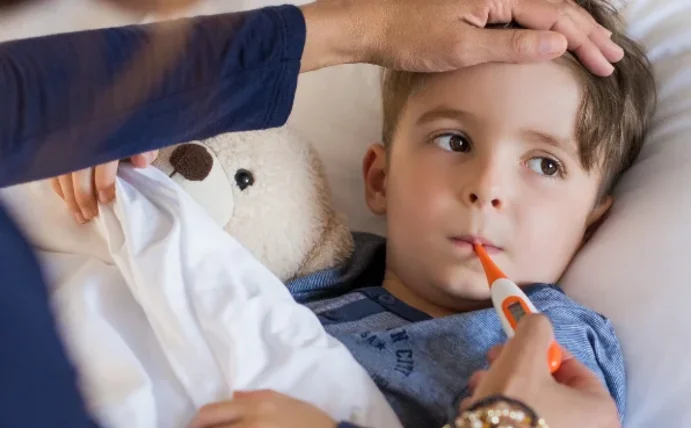 A toddler is lying in bed with a thermometer in their mouth. A woman, probably the mother, is placing a hand on the child’s forehead. 