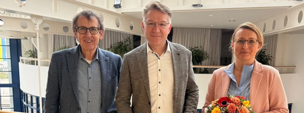Drei Menschen stehen im Hauptgebäude der Uni W/H. In der Mitte steht Prof. Dr. Dr. Lars Bonitz, der mit einem Lächeln seine Berufungsurkunde in der Hand hält.