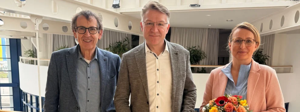 Drei Menschen stehen im Hauptgebäude der Uni W/H. In der Mitte steht Prof. Dr. Dr. Lars Bonitz, der mit einem Lächeln seine Berufungsurkunde in der Hand hält.