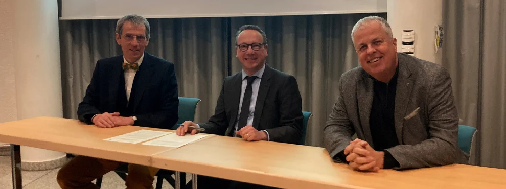 Drei Personen sitzen nebeneinander an einem Tisch und lächeln in die Kamera.