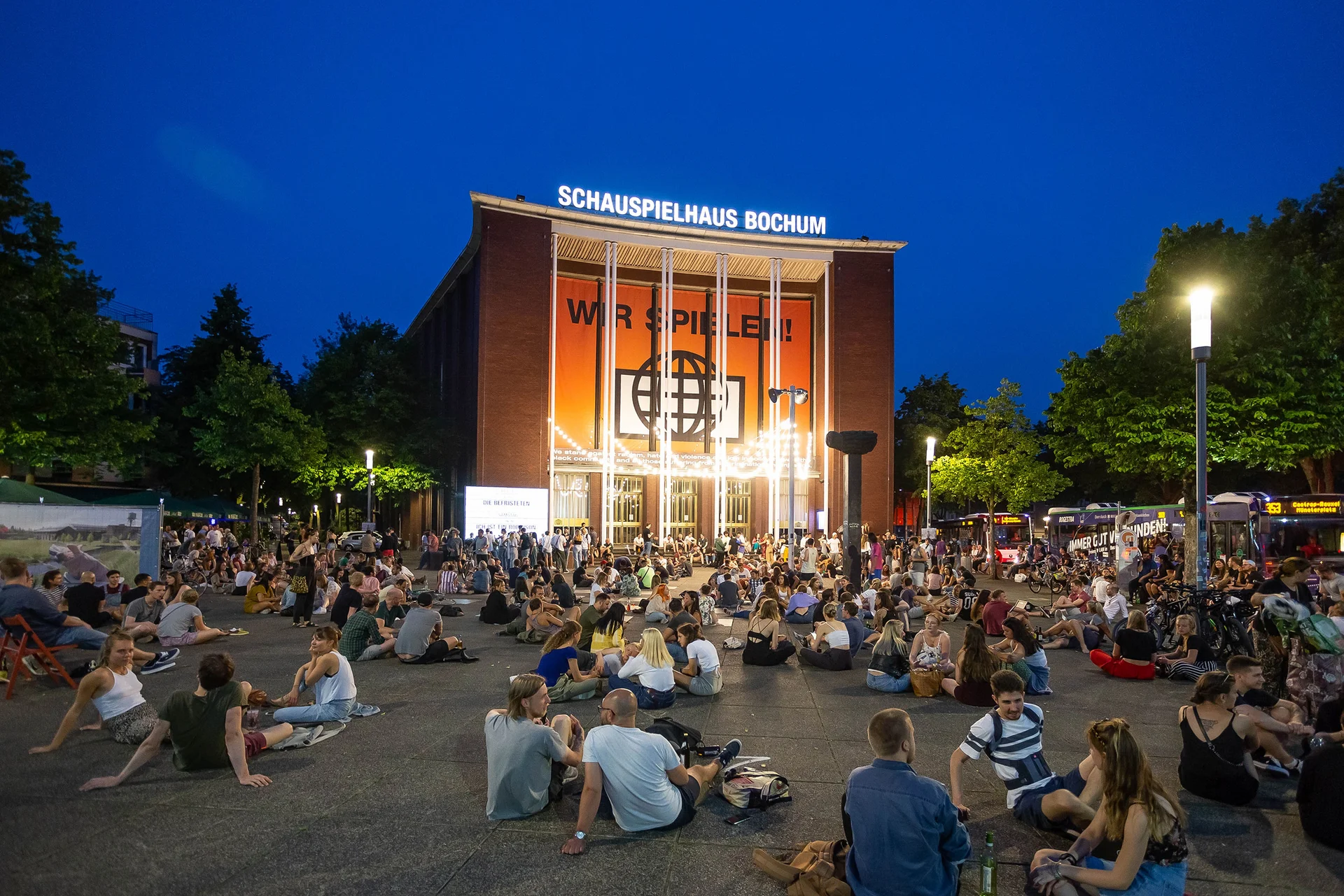 Abendliche Außenaufnahme vom Bochumer Schauspielhaus mit Vorplatz. Hier sitzen viele Menschen in Grüppchen auf dem Boden und unterhalten sich.