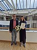 Ein Mann und eine Frau stehen auf vor einer Brüstung der Universität Witten/Herdecke und lächeln in die Kamera. Die Frau hält ihre Berufungsurkunde in der Hand.