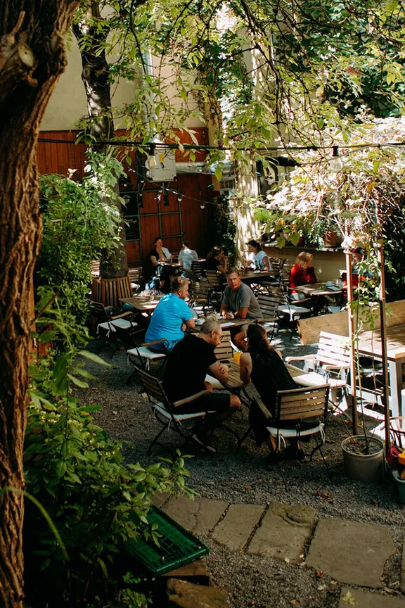 A view of a beer garden: there is a tree at the left-hand edge of the picture. In the centre, people are sitting at a set of wooden tables and chairs.