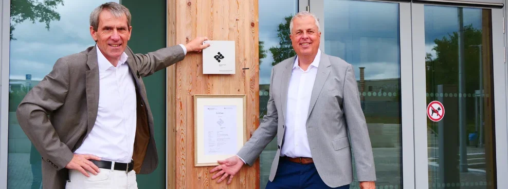 Two men stand in front of a wooden building with a certificate hanging from it and smile into the camera.