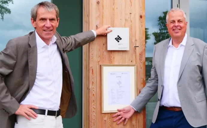 Zwei Männer stehen vor einem Holzgebäude, an dem eine Urkunde hängt, und lächeln in die Kamera.