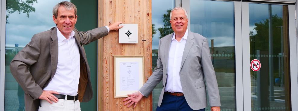 Zwei Männer stehen vor einem Holzgebäude, an dem eine Urkunde hängt, und lächeln in die Kamera.