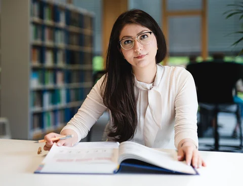 Bewirb dich für den Studiengang Management (B. Sc.) an der UW/H (Foto: UW/H | Johannes Buldmann) Eine junge Frau sitzt in einer Bücherei an einem Tisch mit einem Buch vor sich.