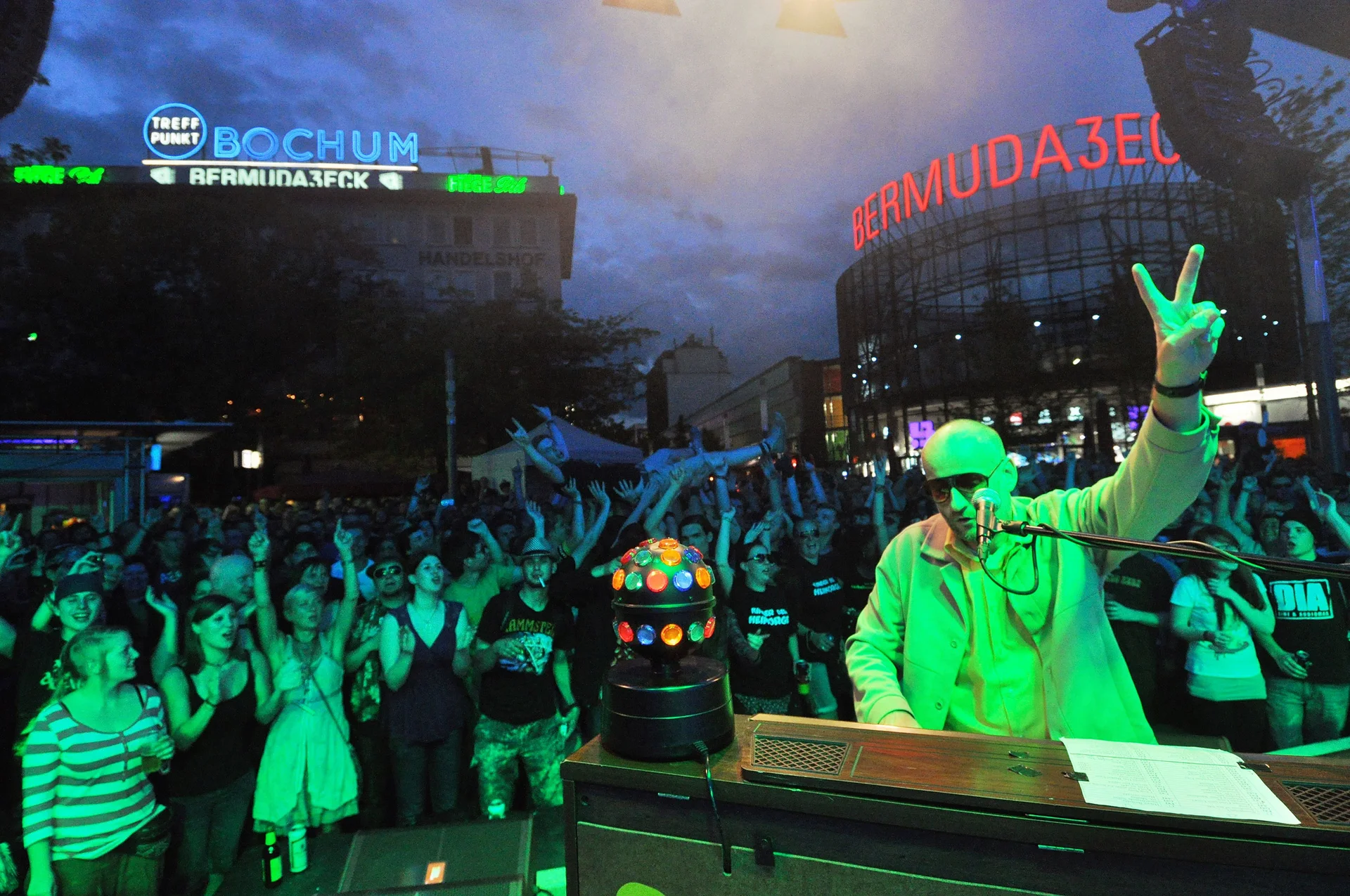 Nightlife at the Bermuda 3Eck in Bochum (Photo: City of Bochum) A night-time shot of the trendy Bermuda 3Eck district. The focus is on a DJ. Lots of people are dancing beside him. The Bermuda 3Eck sign is lit up in the background.