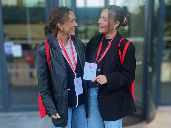 Foto von Clara und Camille vor einer Glastür. Beide lächeln sich an.