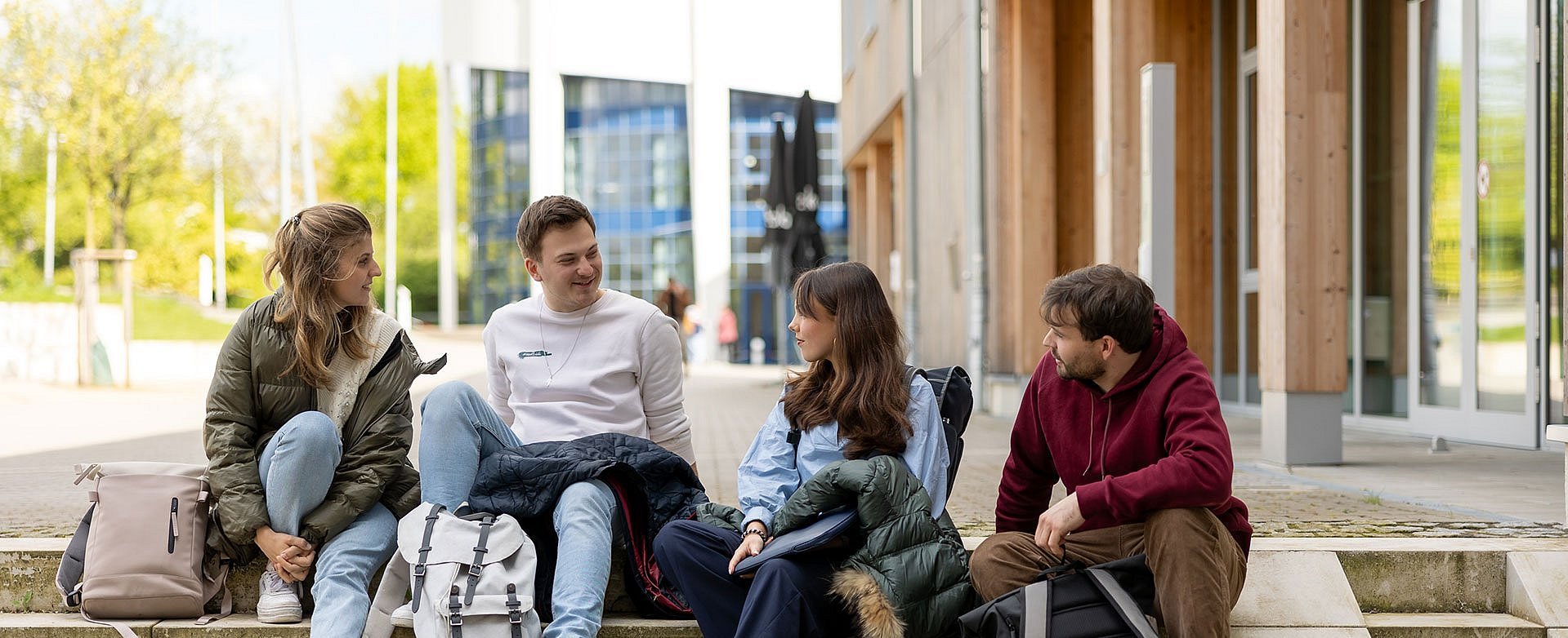 News und Aktuelles aus dem Unileben an der UW/H (Foto: UW/H | Micheal Schwettmann) Vier Studierende sitzen draußen auf einer Treppe vor dem Unigebäude.