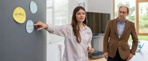 Eine Masterstudentin hält einen Vortrag an einer Tafel, der Professor steht dabei