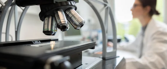 In ihrer Forschung schafft die UW/H Wissen für Mensch und Gesellschaft (Foto: westview | Ingo Beisheim) Nahaufnahme eines Mikroskops im Vordergrund, im Hintergrund unscharf eine Wissenschaftlerin am Schreibtisch