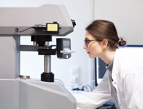 Gesundheitsforschung an der UW/H. (Foto: UW/H | Michael Schmettmann) Zu sehen ist eine Wissenschaftlerin im Profil, die durch ein Mikroskop schaut. Sie trägt eine Brille und einen weißen Laborkittel.