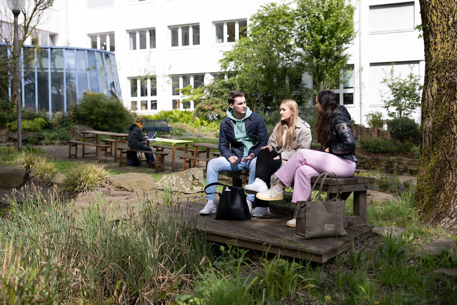 Three students are sitting on a bench in the green university garden. The university building can be seen behind them.