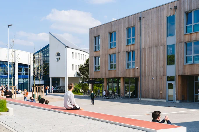 Campus of the UW/H (Photo: UW/H | Michael Schwettmann) Overview of the campus of Witten/Herdecke University. On the left is the white and blue main building, on the right the wooden building. In the foreground, people are sitting on concrete steps.