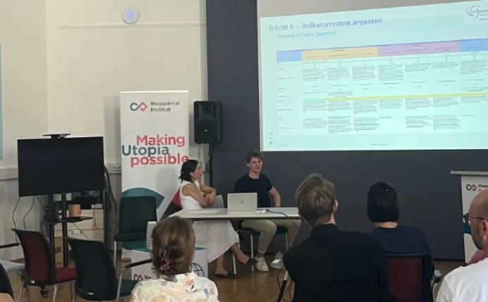 Presentation photo: in the foreground, two students stand in front of a projected PowerPoint presentation; in the background, several people in the audience can be seen from behind 