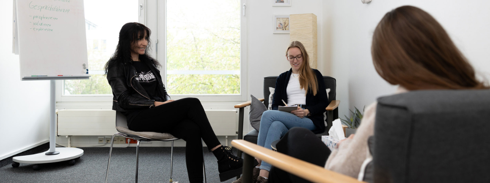 Drei Menschen sitzen in einer Therapiesituation zusammen, zwei davon in Sesseln