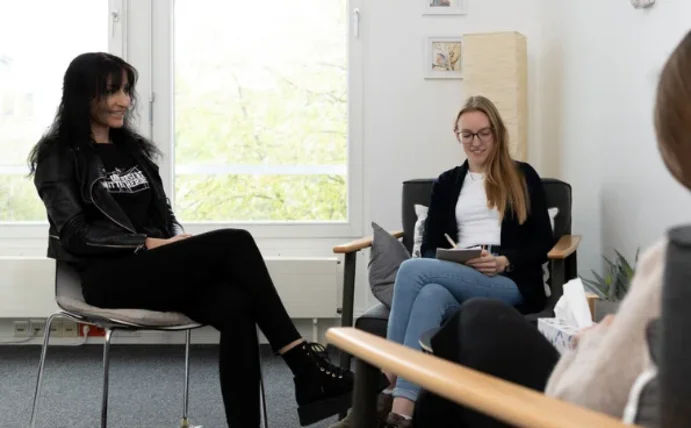 Drei Menschen sitzen in einer Therapiesituation zusammen, zwei davon in Sesseln