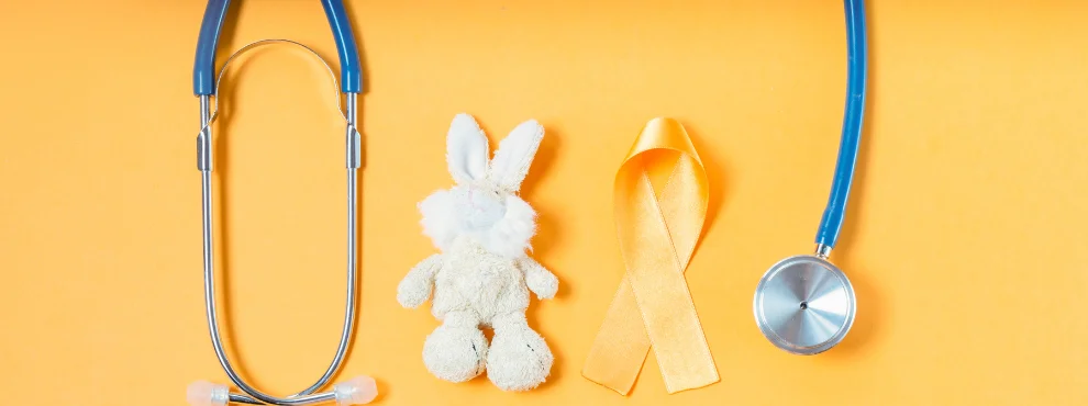 Auf einem gelbgoldenen Hintergrund sieht man ein Stethoskop, eine goldene Schleife und ein Stoffhäschen.