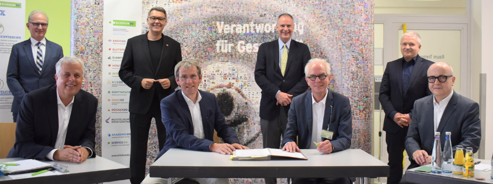 Acht Männer lächeln ins Bild, vier sitzen an einem Tisch im Vordergrund, vier stehen dahinter.