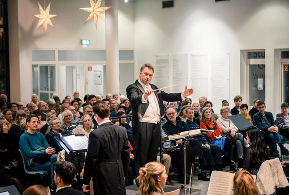 A conductor stands in front of an orchestra. The audience sits in the background.