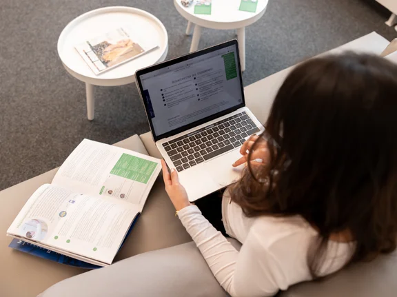 Die Kamera schaut einer Studentin über die Schulter, die auf einem Sofa sitzt. Sie hat ihren Laptop auf dem Schoß; neben ihr liegt eine Broschüre auf dem Sofa. 