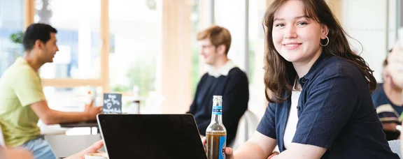 Bewirb dich für den Studiengang PPE – Philosophy, Politics and Economics (M. A.) an der UW/H. (Foto: UW/H | Johannes Buldmann) Eine junge Frau sitzt mit einem Softdrink an einem Tisch vor einem Laptop.