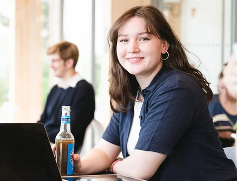 A young woman is sitting at a table in front of a laptop with a soft drink.