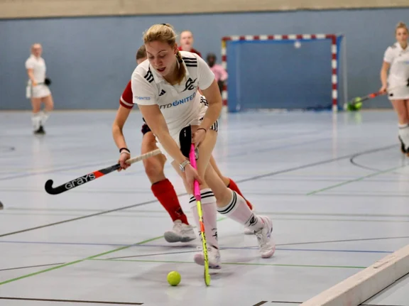 Eine Hockey-Spielerin im Zweikampf um den Ball. Das Spiel findet in einer Sporthalle statt.