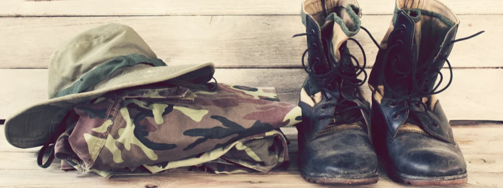 Man sieht die Kleidung eines Soldaten. Eine gefaltete Hose in Tarnfarben, Stiefel und einen Hut.