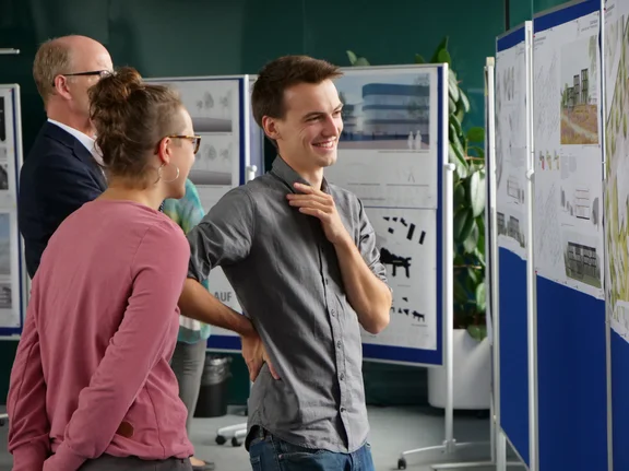Um die Bedürfnisse von Mitarbeitenden und Studierenden abzufragen, hat das Neubau-Team alle eingereichten Entwürfe der Architekturbüros präsentiert und Rückmeldungen ausgewertet. (Foto: UW/H | Johannes Buldmann) Domenik steht mit anderen Mitarbeitenden vor einer Metaplanwand mit Bauplänen