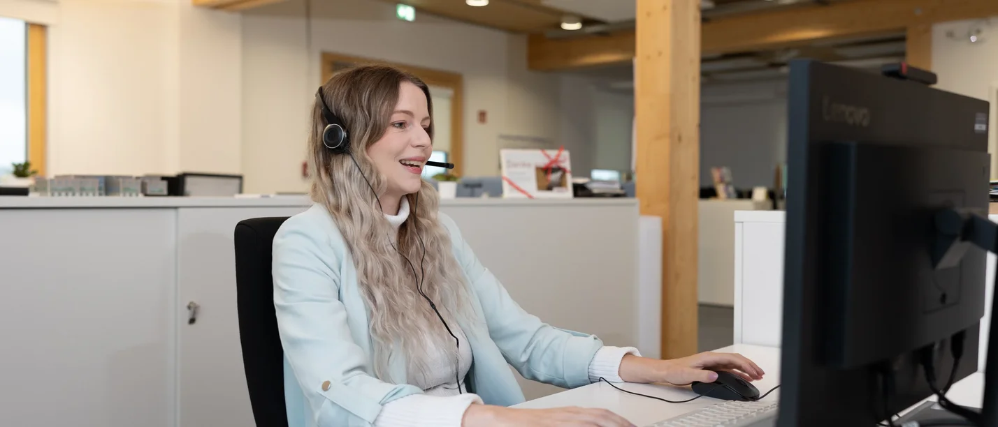 Eine junge Frau mit einem Headset sitzt in einem Großraumbüro vor einen Computer.
