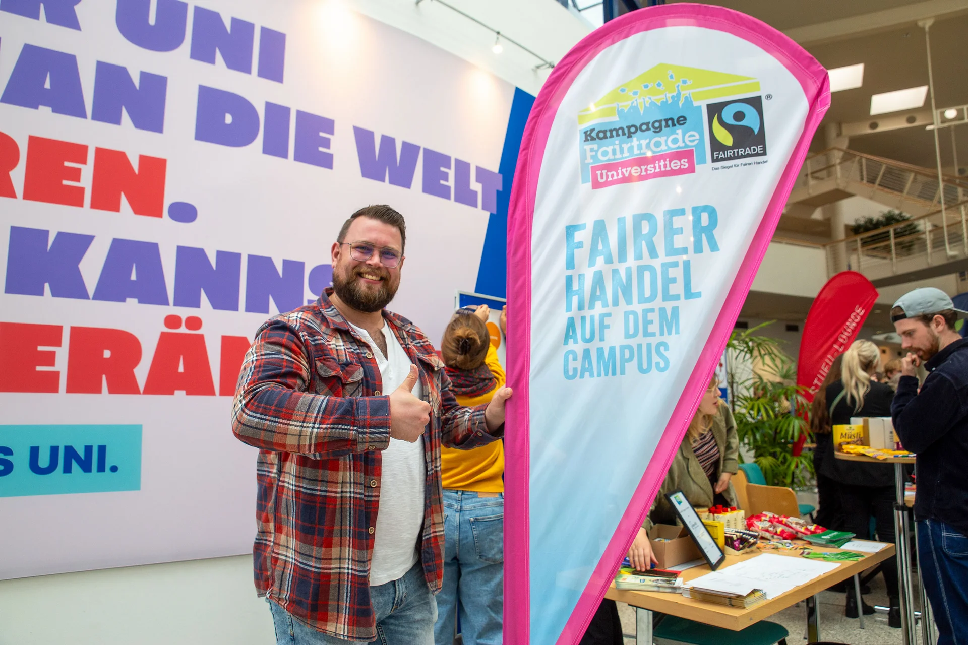 The UW/H is an accredited Fairtrade University. (Photo: UW/H) A man smiles at the camera, giving a thumbs-up. He is standing next to a beach flag bearing the ‘Fairtrade University’ logo.