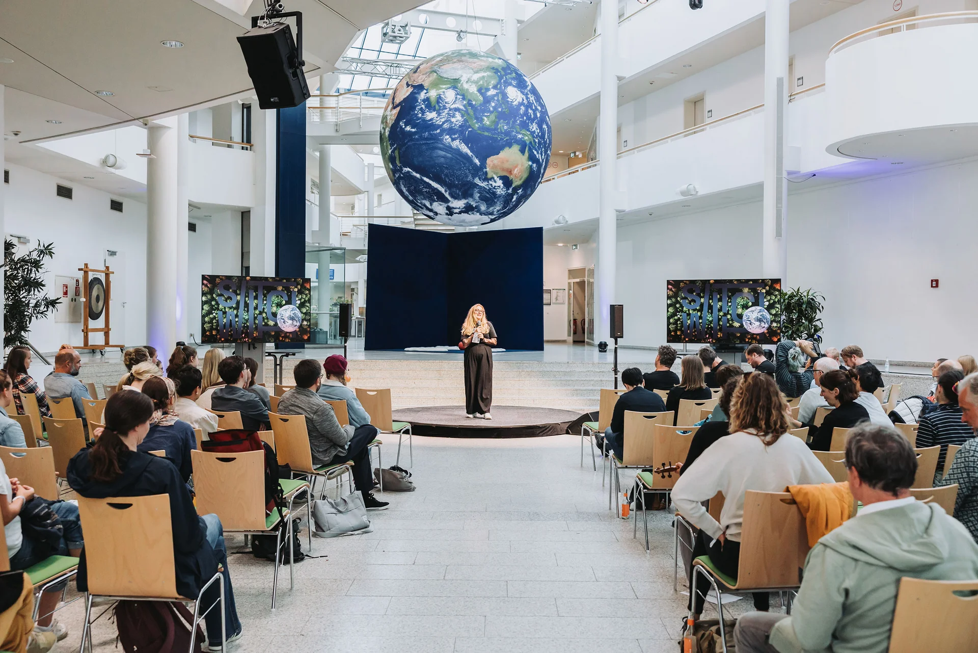 In der Haupthalle der UW/H sitzen Konferenzteilnehmende und blicken zur Rednerin in der Mitte der Halle. Von der Decke hängt eine große Erdkugel.