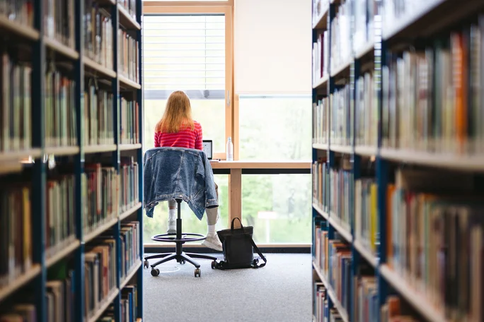 Learning in the library (Photo: UW/H | Johannes Buldmann) The view wanders between two bookshelves to a student who can be seen from behind, sitting at a table and looking towards the window while studying on her laptop.