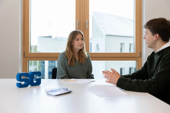 Counselling of the student society (Photo: UW/H | Michael Schwettmann) A woman and a man are sitting opposite each other. There is a large window behind them. The letters "S" and "G" are displayed on the table between them, with documents lying between them.