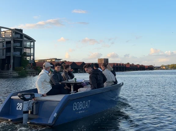 Eine Gruppe von Menschen sitzt in einem Boot auf dem Wasser. Im Hintergrund sind Gebäude.