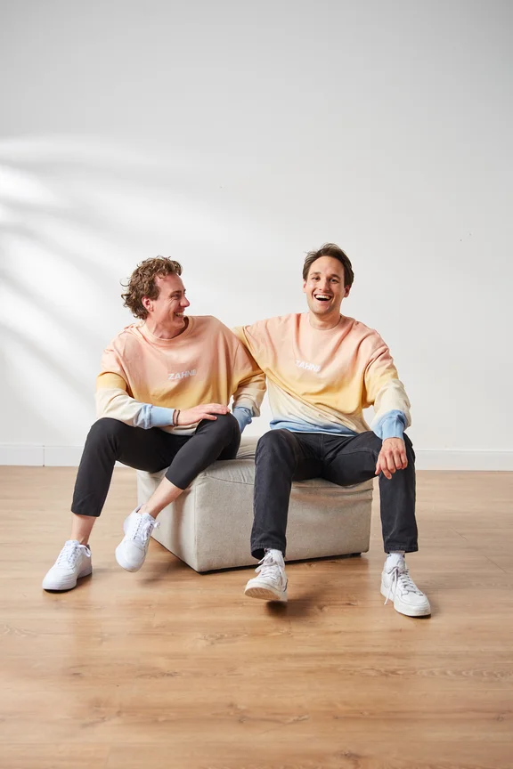 Foto von zwei Männern, die nebeneinander auf einem Hocker sitzen und lachen.