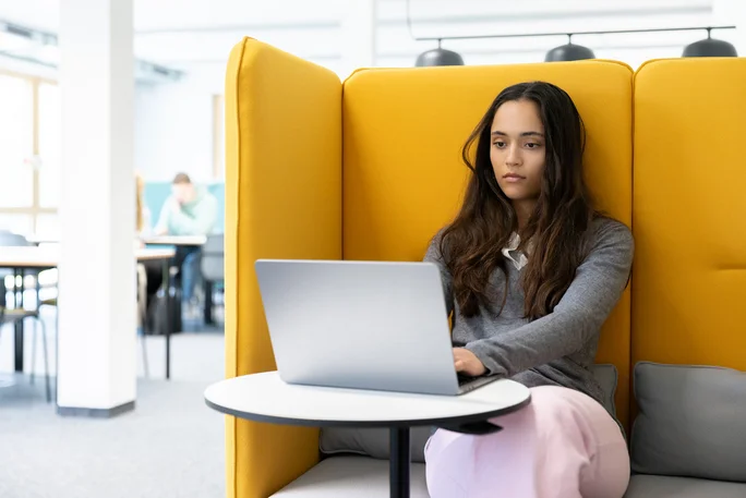 Leseecke in der Bibliothek (Foto: UW/H | Michael Schwettmann) Eine Studentin sitzt in einer gelben Sitzecke in der Bibliothek. Vor ihr steht ihr Laptop auf einem kleinen runden Tisch.