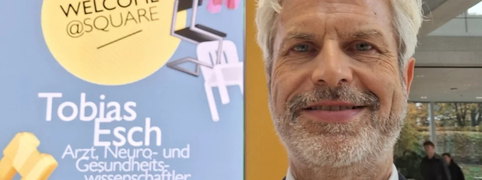 Prof. Dr Tobias Esch stands in front of a programme poster