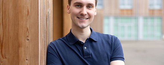 Bewirb dich für den Modellstudiengang Medizin (StEx) an der UW/H. (Foto: UW/H | Michael Schwettmann) Ein junger Mann steht seitlich gelehnt an einer Gebäudewand aus Holz.