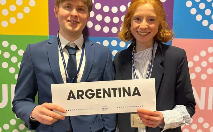 Zwei Studierende stehen vor einer bunten Wand und halten ein Schild mit der Aufschrift "Argentina" hoch.
