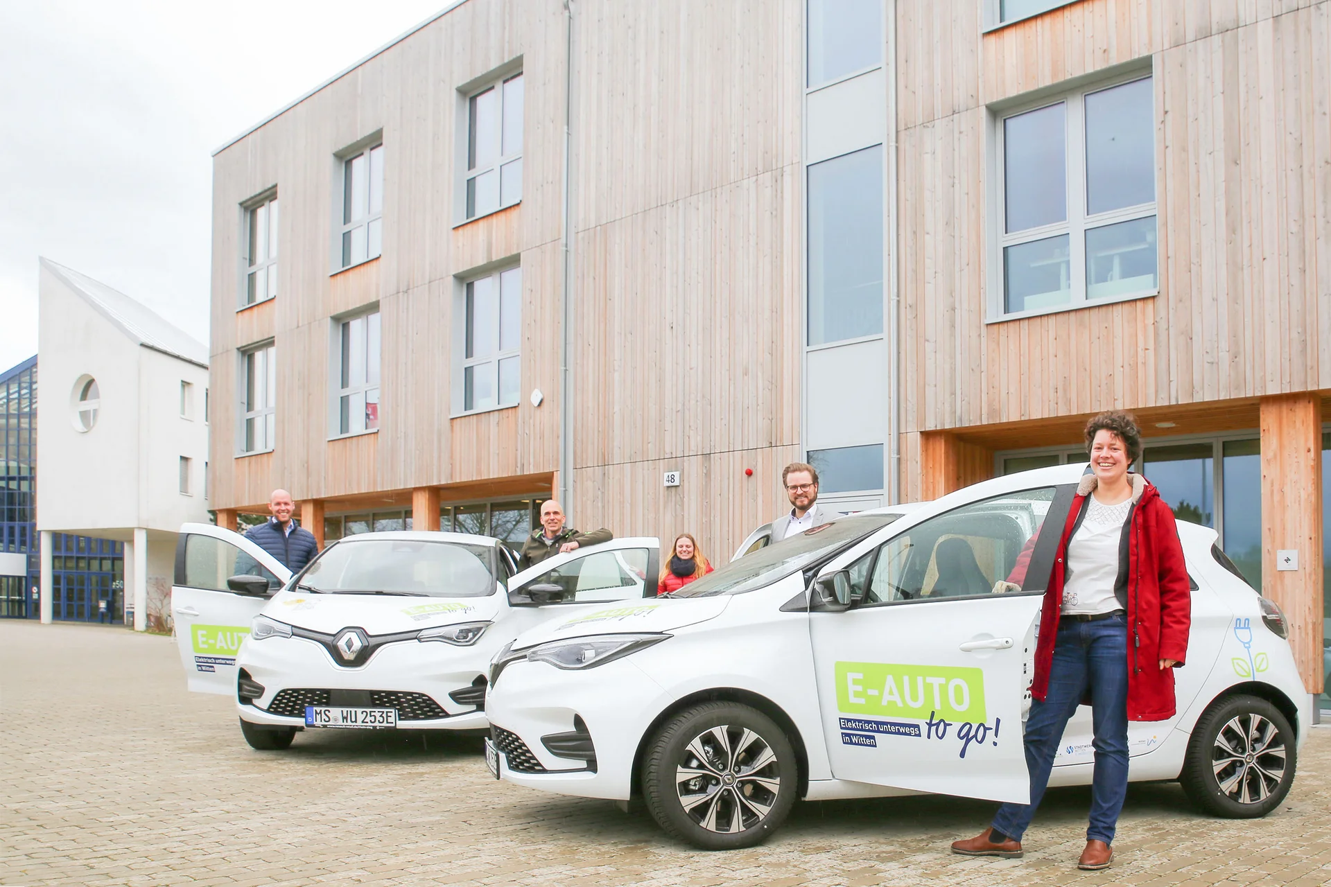 Travelling sustainably with UW/H’s e-car-sharing scheme. (Photo: UW/H | Lucy Mindnich) Two white electric cars with their driver’s doors open are parked in front of the new building. The people in charge of the project are standing next to them.