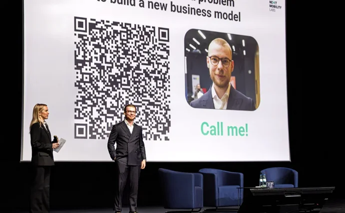 Fabian Schmidt steht auf einer Bühne vor einer weißen Leinwand. Neben ihm steht eine Frau, die ebenfalls moderiert.