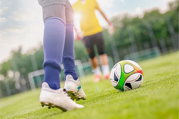 Gemeinsam kicken (Foto: Canva) Im Fokus des Bildes sind die Beinpaare von zwei Fußballspielern. Der vordere von ihnen setzt zum Torschuss an.
