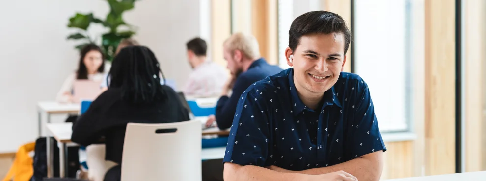 Ein Student sitzt an einem Tisch und lächelt in die Kamera.
