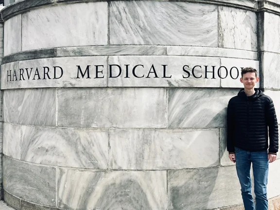 Adrian Illmann, a student, is standing in front of the Harvard Medical School’s building signage and smiling at the camera.