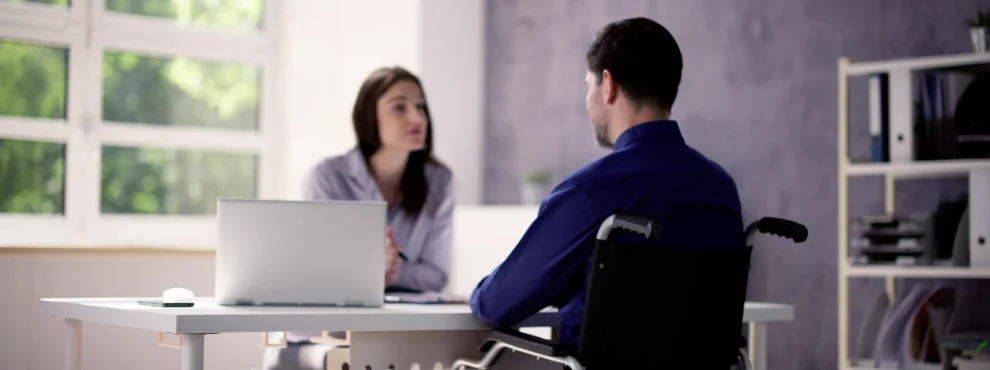 Ein Mann im Rollstuhl sitzt in der Sprechstunde einer Ärztin am Schreibtisch gegenüber.
