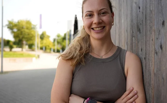 Porträt einer jungen Frau, die mit verschränkten Armen an einer Holzwand lehnt.